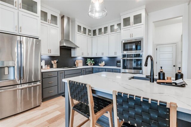 kitchen with gray cabinets, wall chimney exhaust hood, light wood-style flooring, appliances with stainless steel finishes, and backsplash