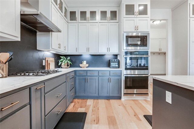 kitchen with decorative backsplash, light wood-style floors, wall chimney exhaust hood, white cabinetry, and stainless steel appliances