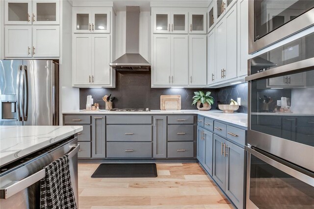 kitchen featuring wall chimney range hood, appliances with stainless steel finishes, light wood-style floors, gray cabinets, and white cabinetry