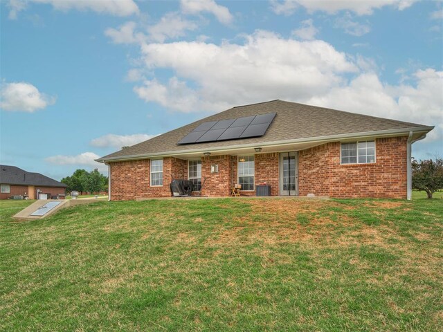 back of property featuring roof with shingles, brick siding, central AC unit, solar panels, and a lawn
