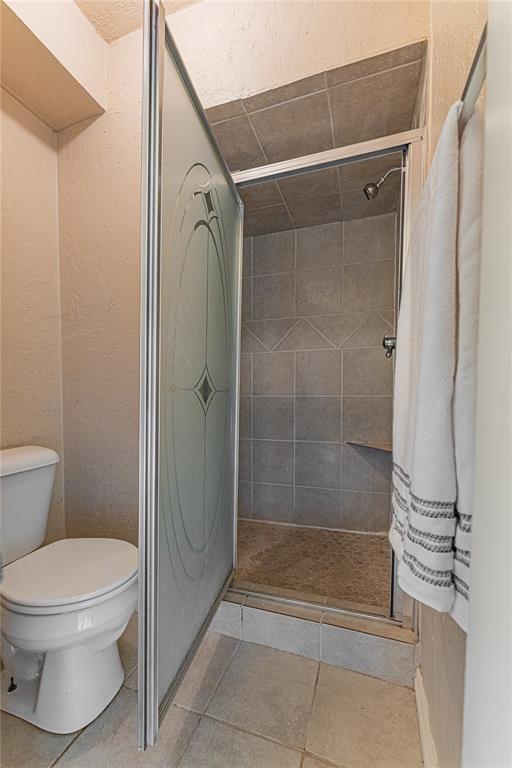 bathroom with toilet, a textured wall, tile patterned floors, and a stall shower