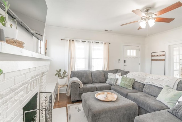 living room featuring a ceiling fan, visible vents, crown molding, a brick fireplace, and wood finished floors