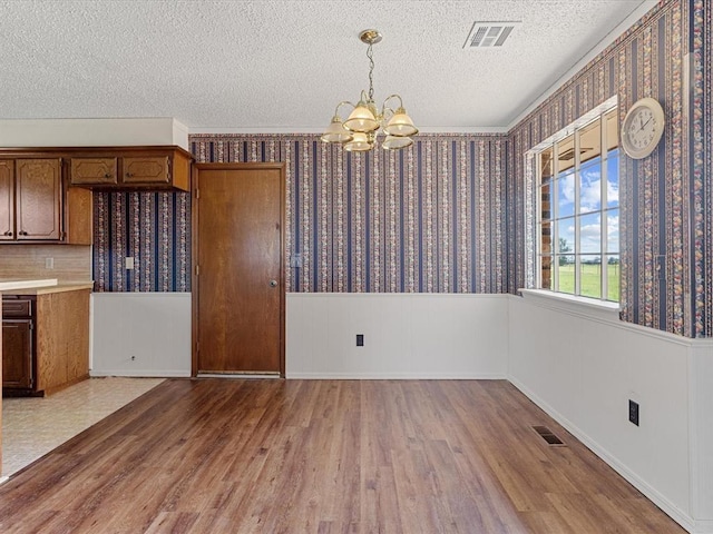 unfurnished dining area with a chandelier, wood finished floors, a textured ceiling, a wainscoted wall, and wallpapered walls