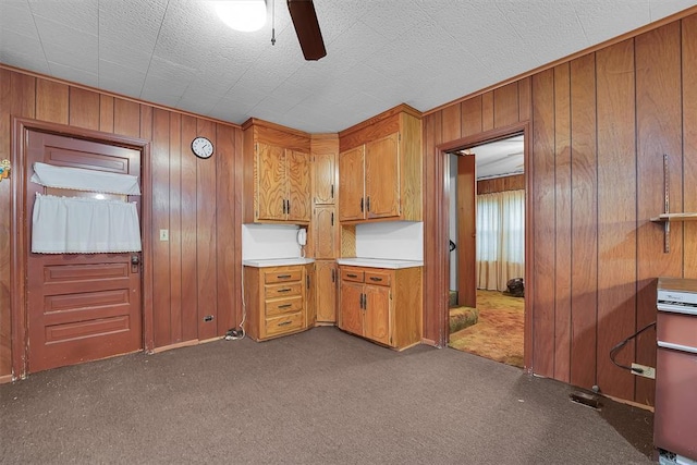kitchen with wooden walls, ceiling fan, light countertops, brown cabinetry, and dark colored carpet