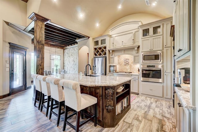 kitchen with open shelves, light stone countertops, an island with sink, stainless steel appliances, and light wood-type flooring