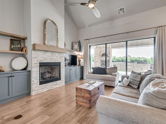 living room featuring visible vents, light wood-style floors, a stone fireplace, ceiling fan, and high vaulted ceiling