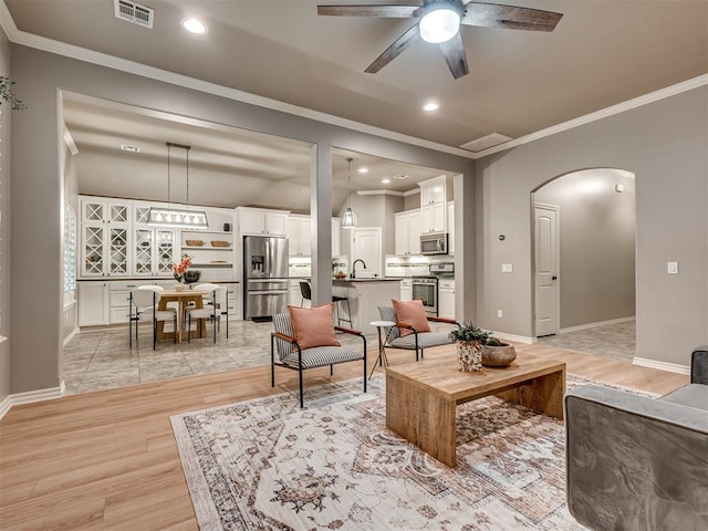 living area featuring ceiling fan, arched walkways, light wood-type flooring, visible vents, and ornamental molding