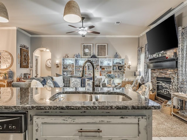 kitchen featuring a sink, a stone fireplace, ceiling fan, arched walkways, and dark stone countertops