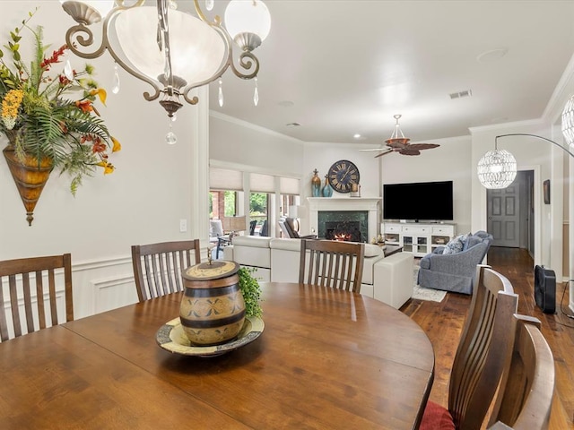 dining space with a ceiling fan, a warm lit fireplace, crown molding, wood finished floors, and a chandelier