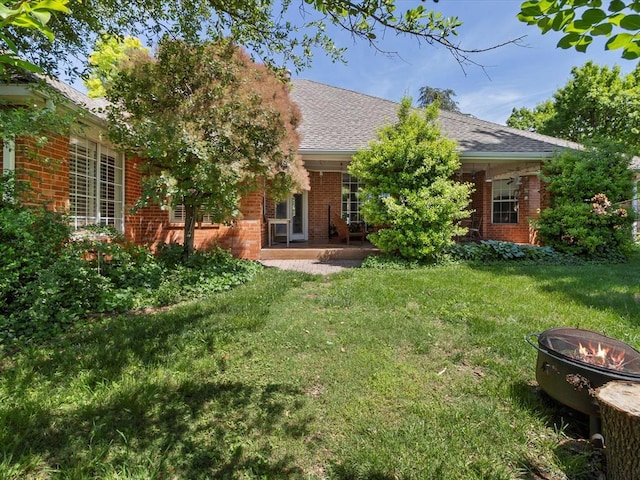 back of property featuring roof with shingles, a patio area, brick siding, an outdoor fire pit, and a yard