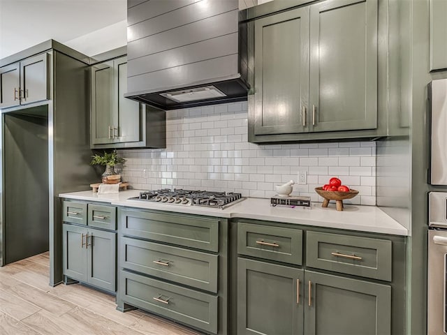 kitchen with wall chimney range hood, backsplash, green cabinetry, and light countertops