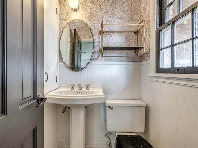 bathroom with toilet, wallpapered walls, wainscoting, and a decorative wall