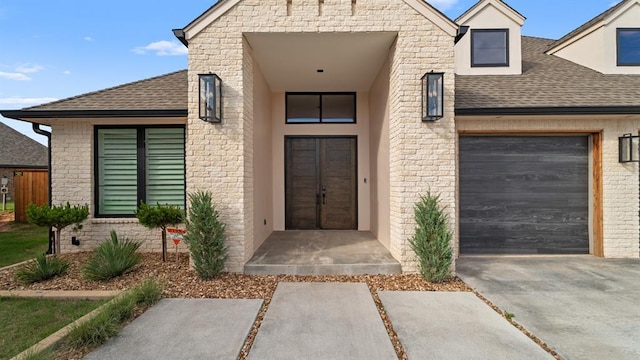 view of exterior entry with roof with shingles, a garage, driveway, and stucco siding