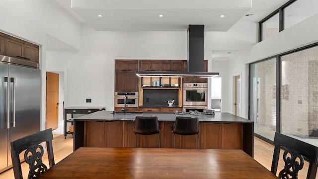 kitchen featuring stainless steel appliances, dark countertops, a high ceiling, light wood-type flooring, and a large island with sink