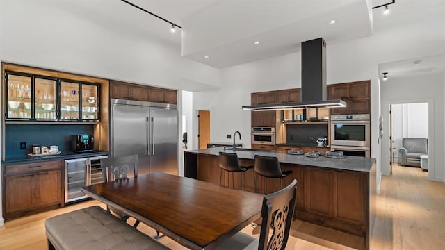 kitchen featuring appliances with stainless steel finishes, rail lighting, beverage cooler, dark countertops, and a center island with sink