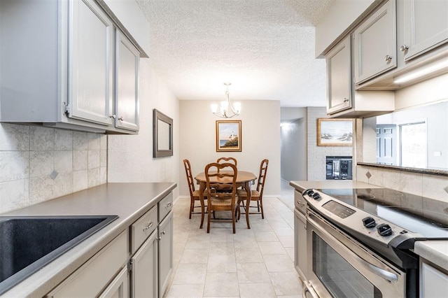 kitchen with stainless steel electric range oven, a notable chandelier, a brick fireplace, gray cabinets, and backsplash