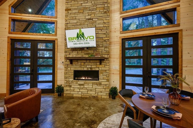 living area featuring french doors, wooden walls, concrete flooring, a stone fireplace, and a towering ceiling