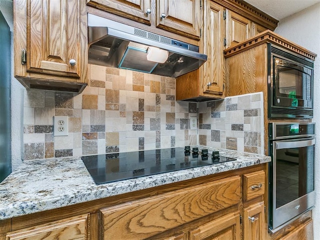 908 Carfax Rd, Edmond, OK 73034 - kitchen with under cabinet range hood, black appliances, backsplash, brown cabinets, and light stone countertops