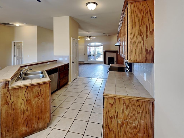 kitchen with a peninsula, a fireplace, tile counters, light tile patterned floors, and stainless steel dishwasher
