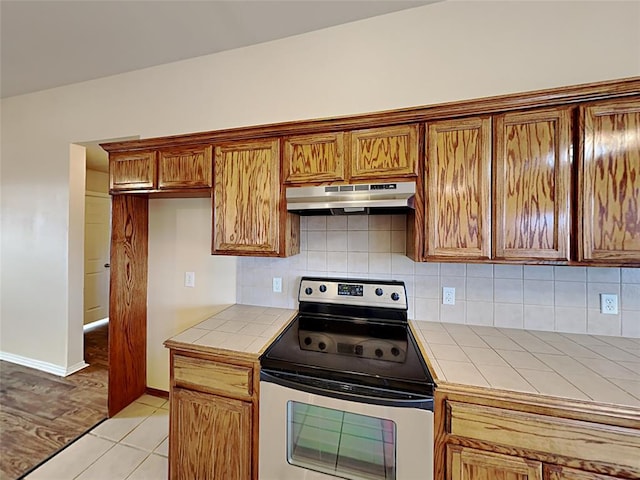 kitchen featuring brown cabinetry, tile countertops, backsplash, under cabinet range hood, and stainless steel electric range oven
