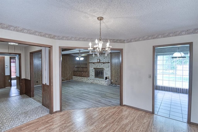 unfurnished dining area with ceiling fan with notable chandelier, wood finished floors, a textured ceiling, a fireplace, and baseboards