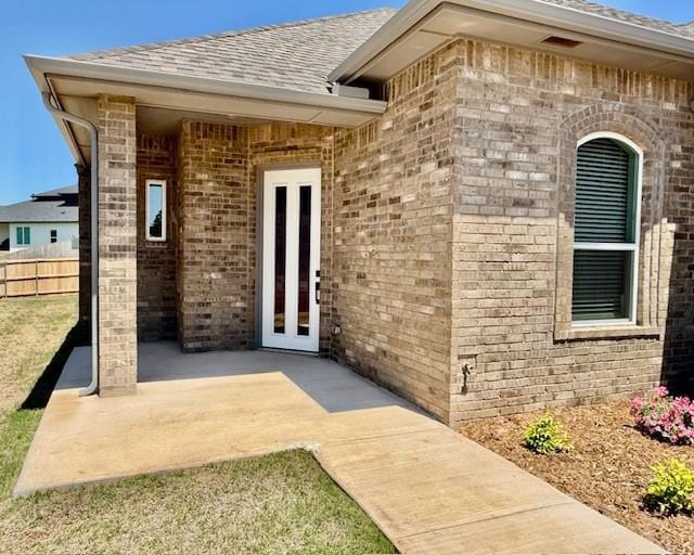 view of exterior entry featuring fence, brick siding, roof with shingles, a patio, and french doors