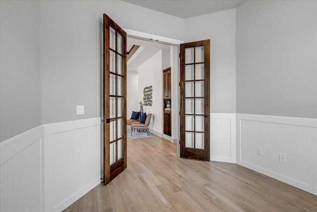 corridor featuring french doors, a wainscoted wall, light wood-style floors, and a decorative wall