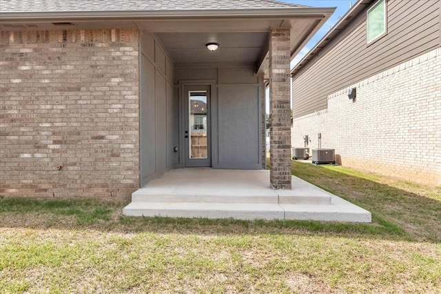 property entrance featuring a yard, roof with shingles, brick siding, and central AC