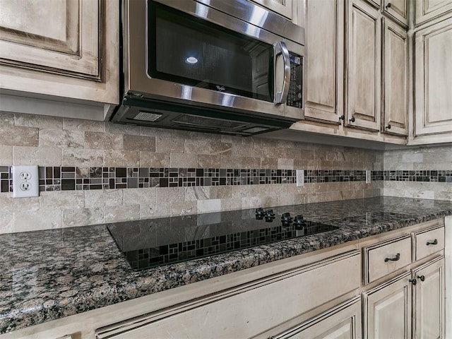 kitchen with stainless steel microwave, backsplash, black electric cooktop, and dark stone countertops