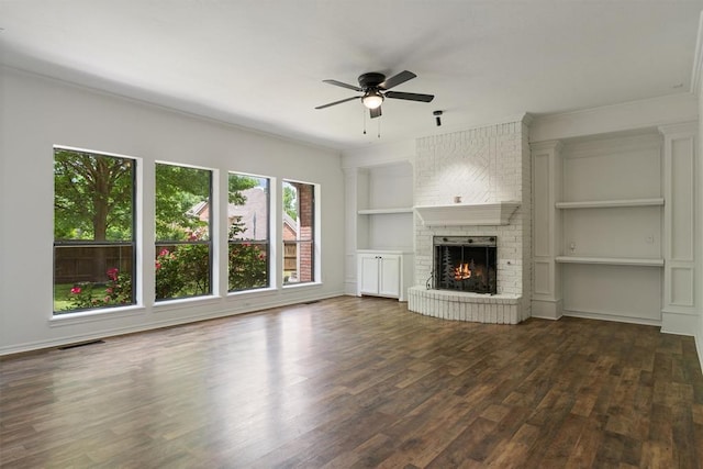 unfurnished living room featuring dark wood-type flooring, built in features, a ceiling fan, a brick fireplace, and ornamental molding