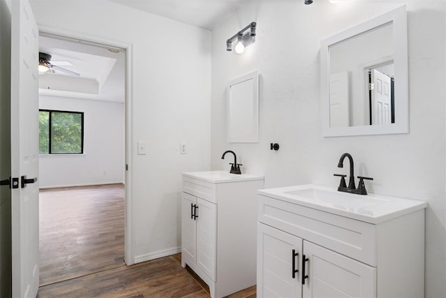 bathroom with two vanities, wood finished floors, a ceiling fan, a tray ceiling, and baseboards