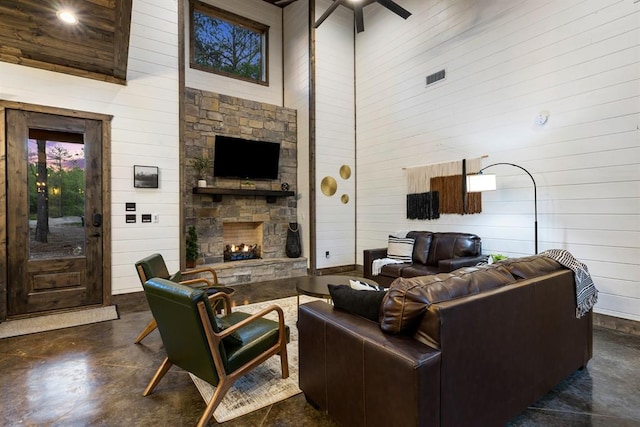 living room featuring a towering ceiling, wooden walls, concrete flooring, a stone fireplace, and a ceiling fan