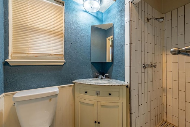 full bathroom with tiled shower, toilet, vanity, wainscoting, and a textured wall