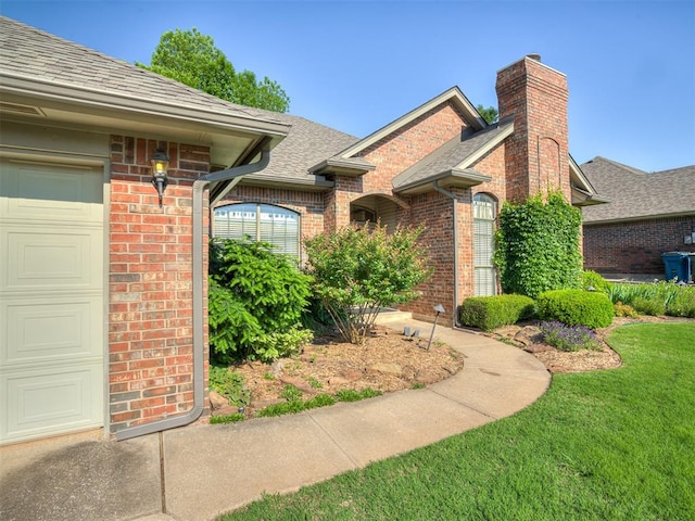 view of side of property with brick siding, an attached garage, a chimney, a shingled roof, and a yard