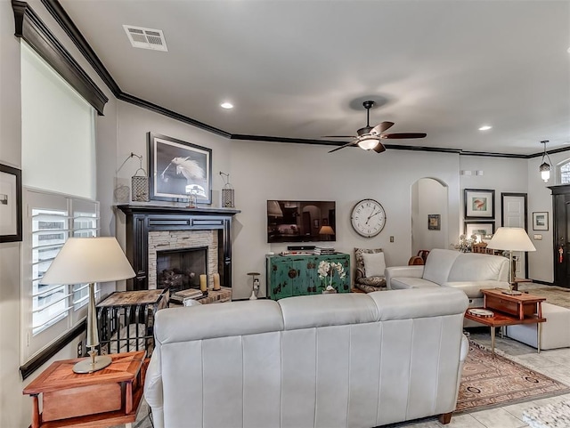 living room with arched walkways, a stone fireplace, ornamental molding, ceiling fan, and recessed lighting