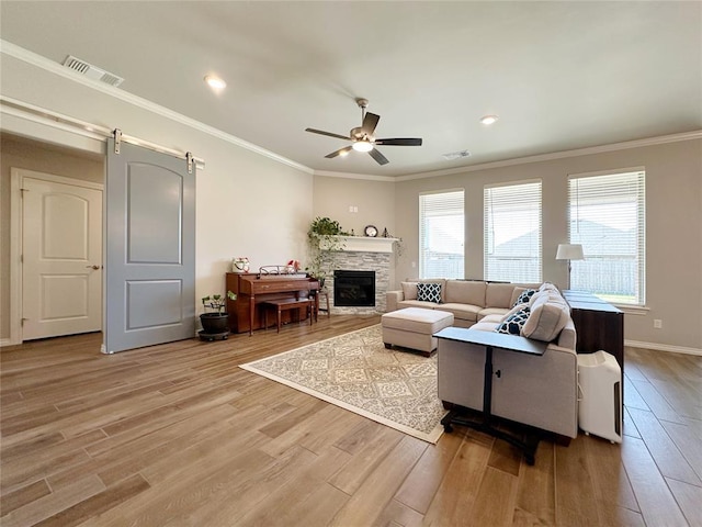 living area featuring light wood-style flooring, ceiling fan, a stone fireplace, crown molding, and a barn door