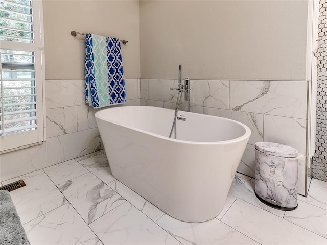 bathroom with tile walls, marble look tile flooring, wainscoting, and a freestanding bath