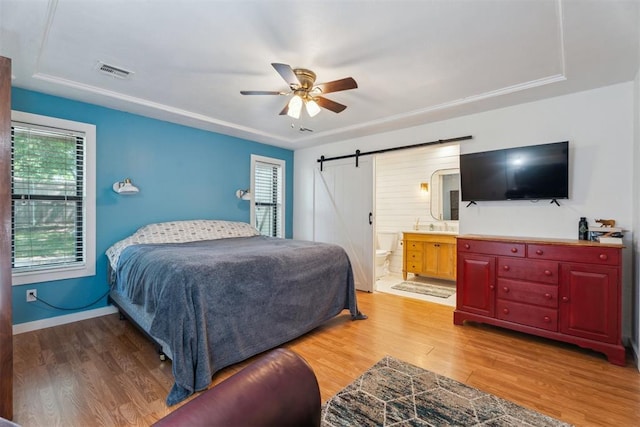 bedroom with light wood-type flooring, a barn door, connected bathroom, a ceiling fan, and baseboards