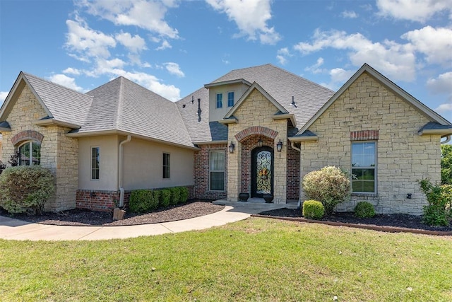 french country inspired facade with stone siding, roof with shingles, a front yard, and stucco siding