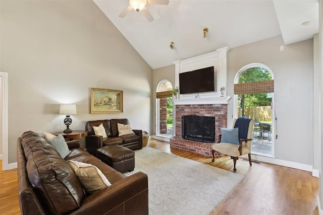living room featuring high vaulted ceiling, a ceiling fan, a fireplace, light wood finished floors, and baseboards