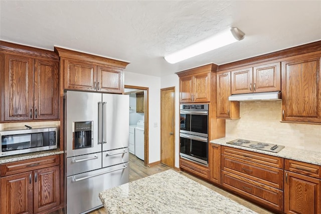 kitchen with appliances with stainless steel finishes, washing machine and clothes dryer, light wood finished floors, backsplash, and under cabinet range hood