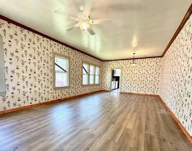 unfurnished living room with wallpapered walls, wood finished floors, crown molding, a ceiling fan, and baseboards