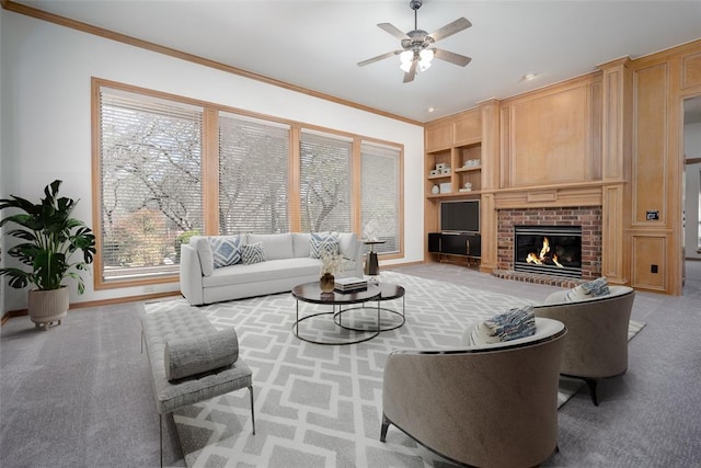 carpeted living area with built in shelves, a ceiling fan, baseboards, a fireplace, and ornamental molding