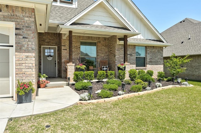 doorway to property featuring a yard, roof with shingles, board and batten siding, and brick siding