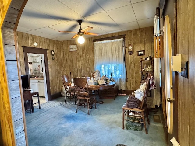 carpeted dining space featuring wood walls, ceiling fan, and a drop ceiling