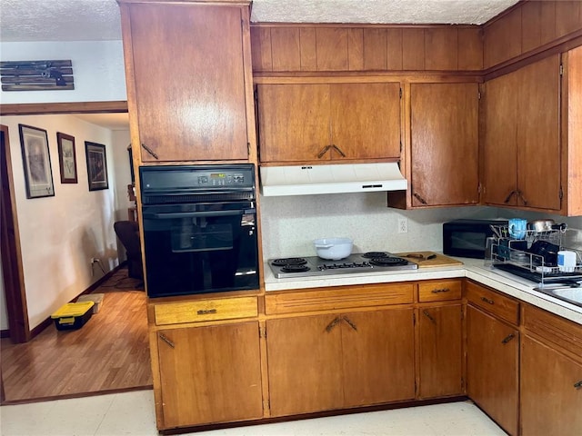 321 E Ninth St, Leedey, OK 73654 - kitchen featuring black appliances, under cabinet range hood, light countertops, brown cabinetry, and a textured ceiling