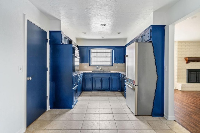 kitchen featuring freestanding refrigerator, blue cabinetry, a sink, a textured ceiling, and a brick fireplace