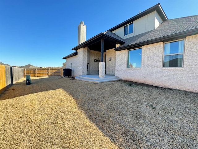 back of house featuring a chimney, a fenced backyard, a shingled roof, central AC, and brick siding
