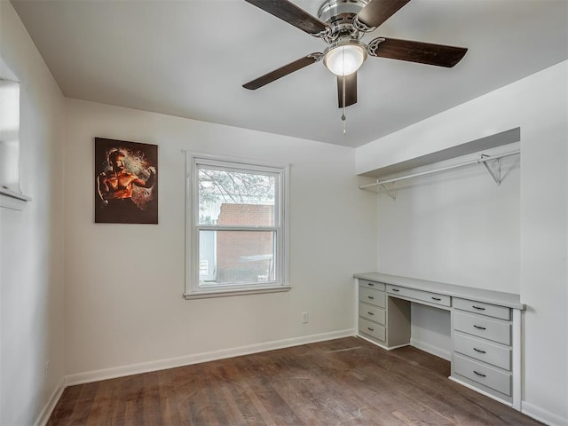 unfurnished office featuring dark wood-type flooring, built in desk, a ceiling fan, and baseboards