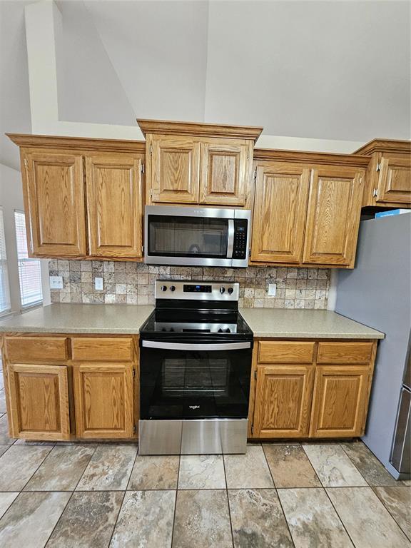 1801 Caribou St, Altus, OK 73521 - kitchen featuring appliances with stainless steel finishes, backsplash, light countertops, vaulted ceiling, and brown cabinets